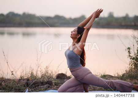 Woman performing a low lunge yoga pose with arms raised beside a lake, showing outdoor wellness, hip flexibility, strength, balance, mindful breathing, mobility, and healthy lifestyle. Woman performing a low lunge yoga pose with arms raised beside a lake, showing outdoor wellness, hip flexibility, strength, balance, mindful breathing, mobility, and healthy lifestyle. 140447738