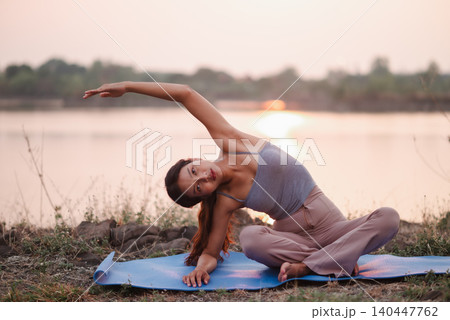Woman doing a seated side bend on a blue yoga mat beside a lake, emphasizing outdoor wellness, flexibility, core strength, mobility, mindful stretching, calm balance, and healthy lifestyle. Woman doing a seated side bend on a blue yoga mat beside a lake, emphasizing outdoor wellness, flexibility, core strength, mobility, mindful stretching, calm balance, and healthy lifestyle. 140447762