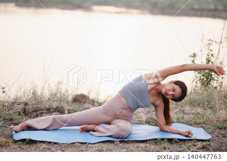 Woman performing a side body yoga stretch on a blue mat by the lake, emphasizing flexibility, core strength, balance, mobility, outdoor fitness, wellness routine, and mindful movement. 140447763