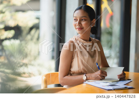 Businesswoman writing notes in a notebook at a modern office desk by large windows, showing planning, productivity, paperwork, financial review, focus, organization, and professional work routine. Businesswoman writing notes in a notebook at a modern office desk by large windows, showing planning, productivity, paperwork, financial review, focus, organization, and professional work routine. 140447828