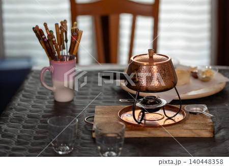 Copper fondue pot with an elegant stand boiling oil on a dining table, ready for cooking various meats with colorful skewers for a gourmet meal experience Copper fondue pot with an elegant stand boiling oil on a dining table, ready for cooking various meats with colorful skewers for a gourmet meal experience 140448358