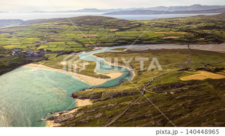 Aerial view shows Bay, estuary, and lush green fields in Ireland. A winding river connects a sandy beach to the sea, observed on a bright, clear day. Aerial view shows Bay, estuary, and lush green fields in Ireland. A winding river connects a sandy beach to the sea, observed on a bright, clear day. 140448965