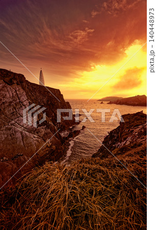 A white lighthouse stands atop rugged cliffs as orange and yellow sunlight paints the sky above the vast ocean. Waves crash below, and dry grass fills the foreground. A white lighthouse stands atop rugged cliffs as orange and yellow sunlight paints the sky above the vast ocean. Waves crash below, and dry grass fills the foreground. 140448973