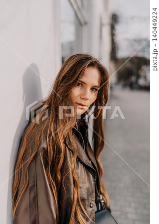 Woman leather jacket street style portrait of a young brunette leaning against a white wall in soft sunlight Woman leather jacket street style portrait of a young brunette leaning against a white wall in soft sunlight 140449224