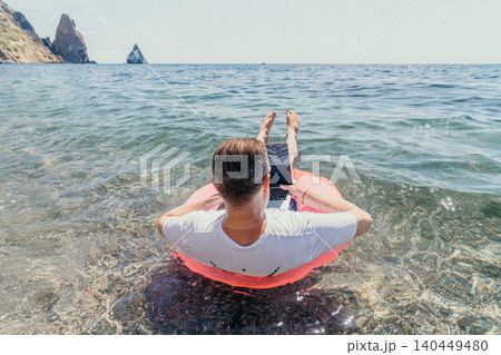 Freelancer laptop sea man floating on a pink inflatable ring while working on a laptop in clear ocean water near rocky cliffs 140449480