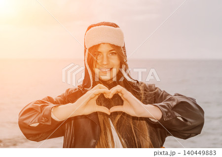 Woman heart gesture young female wearing ushanka makes love sign with hands at sunset sea shore with copy space Woman heart gesture young female wearing ushanka makes love sign with hands at sunset sea shore with copy space 140449883