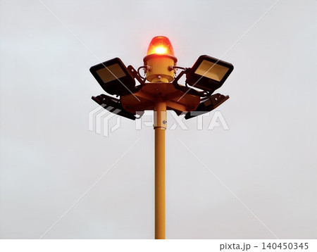Yellow airport apron light mast with red rotating beacon and floodlights against gray sky 140450345