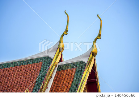 Close up of golden Chofah on traditional Thai temple roof gable with colorful tiles against blue sky background for cultural travel or Asian architectural design concept Close up of golden Chofah on traditional Thai temple roof gable with colorful tiles against blue sky background for cultural travel or Asian architectural design concept 140450360