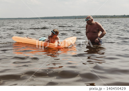 grandfather and grandson enjoy calm summer day. elderly man stands in water holding his child orange inflatable mattress. celebration intergenerational unity, outdoor recreation leisure Family values grandfather and grandson enjoy calm summer day. elderly man stands in water holding his child orange inflatable mattress. celebration intergenerational unity, outdoor recreation leisure Family values 140450429