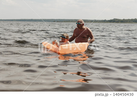 grandfather and grandson enjoy calm summer day. elderly man stands in water holding his child orange inflatable mattress. celebration intergenerational unity, outdoor recreation leisure Family values 140450430