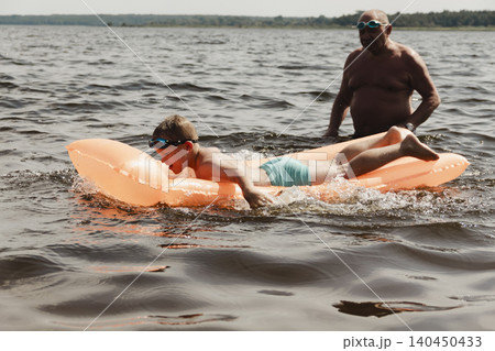 grandfather and grandson enjoy calm summer day. elderly man stands in water holding his child orange inflatable mattress. celebration intergenerational unity, outdoor recreation leisure Family values 140450433