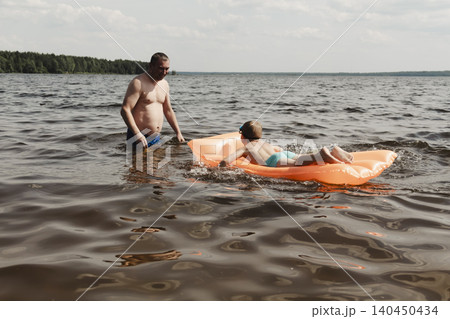 father son enjoying relaxing summer day, floating inflatable mattress calm river. man holding child securely ensuring safety while both enjoy water. Outdoor recreation, family vacation, summer nature father son enjoying relaxing summer day, floating inflatable mattress calm river. man holding child securely ensuring safety while both enjoy water. Outdoor recreation, family vacation, summer nature 140450434