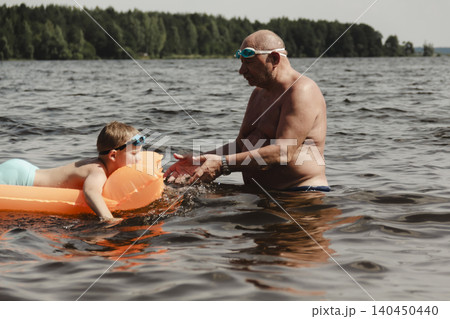 grandfather and grandson enjoy calm summer day. elderly man stands in water holding his child orange inflatable mattress. celebration intergenerational unity, outdoor recreation leisure Family values grandfather and grandson enjoy calm summer day. elderly man stands in water holding his child orange inflatable mattress. celebration intergenerational unity, outdoor recreation leisure Family values 140450440