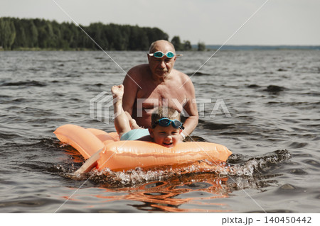 grandfather and grandson enjoy calm summer day. elderly man stands in water holding his child orange inflatable mattress. celebration intergenerational unity, outdoor recreation leisure Family values grandfather and grandson enjoy calm summer day. elderly man stands in water holding his child orange inflatable mattress. celebration intergenerational unity, outdoor recreation leisure Family values 140450442