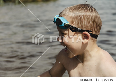 A side profile shot of a young boy in a natural water setting. He is wearing blue swimming goggles and is looking off to the side, seemingly distracted or observing something out of frame A side profile shot of a young boy in a natural water setting. He is wearing blue swimming goggles and is looking off to the side, seemingly distracted or observing something out of frame 140450449