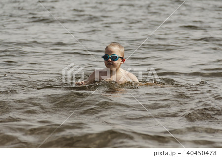 A candid shot of a 6 year old boy swimming in a natural lake. The young swimmer wears dark swimming goggles to protect his eyes from the water and sunlight. Healthy recreation and parenting. A candid shot of a 6 year old boy swimming in a natural lake. The young swimmer wears dark swimming goggles to protect his eyes from the water and sunlight. Healthy recreation and parenting. 140450478
