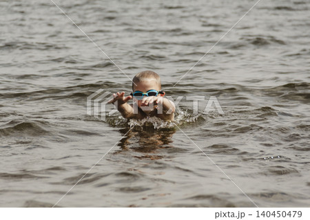 A candid shot of a 6 year old boy swimming in a natural lake. The young swimmer wears dark swimming goggles to protect his eyes from the water and sunlight. Healthy recreation and parenting. A candid shot of a 6 year old boy swimming in a natural lake. The young swimmer wears dark swimming goggles to protect his eyes from the water and sunlight. Healthy recreation and parenting. 140450479