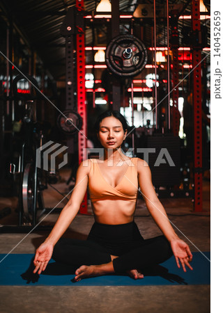 Athletic Asian woman meditating on a blue yoga mat in a dark gym, sitting cross-legged with eyes closed, showing fitness mindfulness, breathing practice, wellness balance, and calm recovery. 140452659