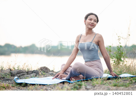 Asian woman sitting on a blue yoga mat beside a quiet lake, resting after practice with a gentle smile, showing outdoor fitness, mindful stretching, relaxation, nature wellness, and peaceful balance. 140452801