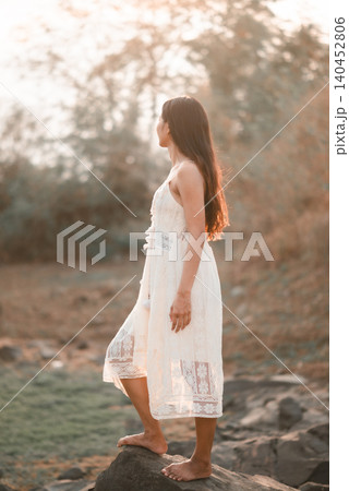Young Asian woman in a white dress standing barefoot on a rock in warm sunset light, looking into nature, showing freedom, confidence, travel escape, serenity, outdoor wellness, and calm lifestyle. Young Asian woman in a white dress standing barefoot on a rock in warm sunset light, looking into nature, showing freedom, confidence, travel escape, serenity, outdoor wellness, and calm lifestyle. 140452806