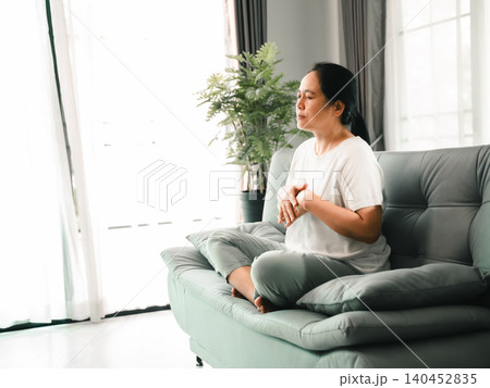 Asian woman meditating cross-legged on a sofa in a bright living room, eyes closed with hands near her chest, showing mindfulness, calm breathing, home wellness, stress relief, relaxation, Asian woman meditating cross-legged on a sofa in a bright living room, eyes closed with hands near her chest, showing mindfulness, calm breathing, home wellness, stress relief, relaxation, 140452835