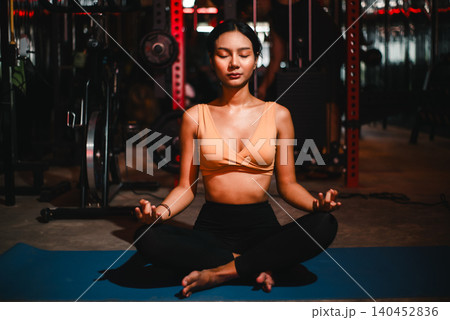 Young athletic Asian woman meditating cross-legged on yoga mat inside a dark gym with eyes closed, showing mindfulness, fitness recovery, balance, breathing, wellness, calm focus, 140452836