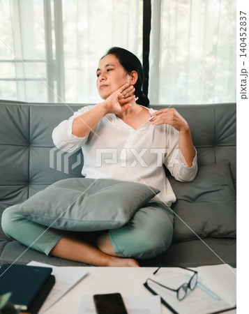 Asian woman sitting on a sofa at home with neck discomfort, touching her shoulder near paperwork and glasses, showing office fatigue, poor posture, stress relief, wellness, body care, 140452837