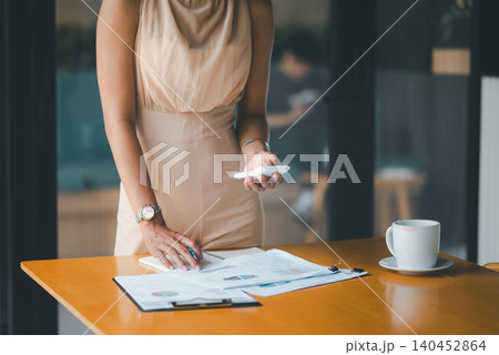 Asian businesswoman checking financial reports on wooden cafe table with smartphone, calculator, notebook, and coffee, showing budget planning, accounting workflow, data review, and corporate finance. 140452864