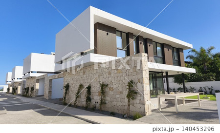 Contemporary luxury villas with clean geometric architecture, stone facades, and large windows under a clear blue sky, showcasing modern residential design and upscale living. 140453296