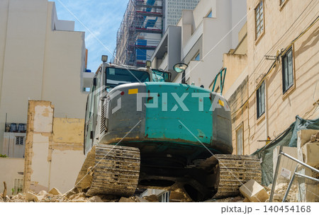 Heavy tracked excavator parked on rubble at urban demolition and construction site surrounded by buildings and scaffolding, industrial machinery photograph.. High quality photo Heavy tracked excavator parked on rubble at urban demolition and construction site surrounded by buildings and scaffolding, industrial machinery photograph.. High quality photo 140454168