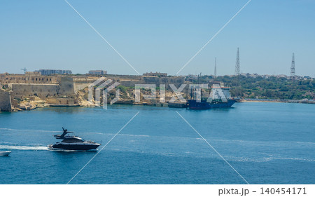 Mediterranean harbor scene with luxury motor yacht, anchored cargo vessel, historic limestone fort and calm blue sea under clear sky in Valletta Malta High quality photo Mediterranean harbor scene with luxury motor yacht, anchored cargo vessel, historic limestone fort and calm blue sea under clear sky in Valletta Malta High quality photo 140454171