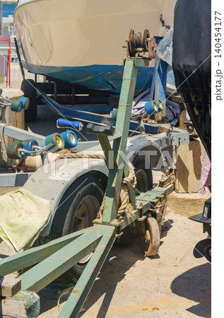 Boats parked on rusty steel trailers with blue rollers, manual winch and mooring ropes at a Mediterranean quayside boat storage yard under bright sun High quality photo 140454177