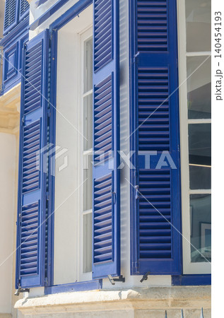 Traditional blue wooden window shutters on white Mediterranean house facade in sunlight, classic European coastal architecture Traditional blue wooden window shutters on white Mediterranean house facade in sunlight, classic European coastal architecture 140454193