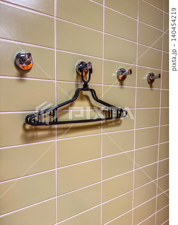 Black plastic clothes hanger hanging on a chrome hook against a olive green ceramic tiled wall in a bathroom or cloakroom. 140454219