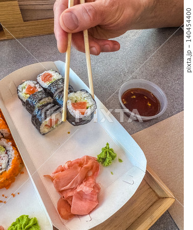 Hand holding salmon maki roll with wooden chopsticks over takeaway box. Dipping sushi in soy sauce container close up. Hand holding salmon maki roll with wooden chopsticks over takeaway box. Dipping sushi in soy sauce container close up. 140454400