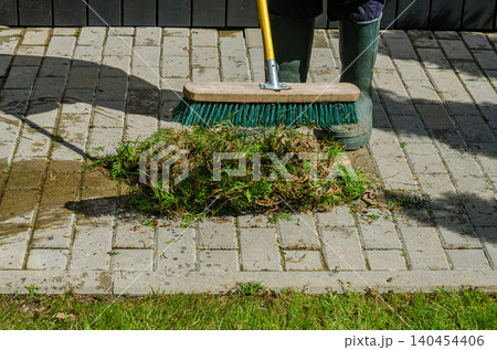 Gardener in Green Rubber Boots Sweeping Conifer Clippings on Stone Paved Walkway with Industrial Turquoise Push Broom Outdoors Gardener in Green Rubber Boots Sweeping Conifer Clippings on Stone Paved Walkway with Industrial Turquoise Push Broom Outdoors 140454406