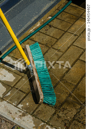 Outdoor Cleaning Concept: Turquoise Industrial Broom Sweeping Wet Stone Pavement Tiles with Garden Hose in the Background Outdoor Cleaning Concept: Turquoise Industrial Broom Sweeping Wet Stone Pavement Tiles with Garden Hose in the Background 140454407