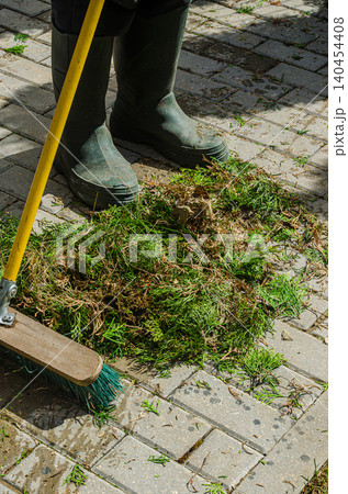 Garden Maintenance and Cleanup: Person in Green Rubber Boots Sweeping Conifer Clippings on Stone Pavement with Outdoor Push Broom 140454408