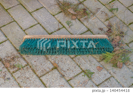 A wooden push broom with green plastic bristles lies flat on outdoor patio pavers amidst scattered dry grass, dead leaves, and yard maintenance debris.. High quality photo A wooden push broom with green plastic bristles lies flat on outdoor patio pavers amidst scattered dry grass, dead leaves, and yard maintenance debris.. High quality photo 140454418