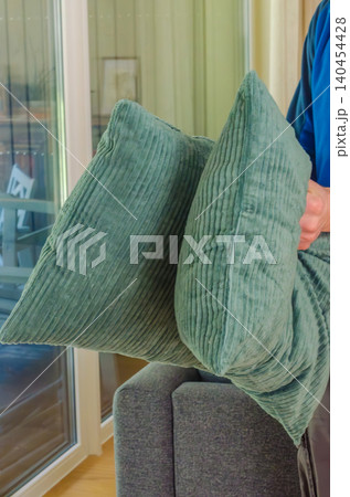 Close-up view of someone holding two textured green corduroy throw pillows indoors near a grey sofa and bright glass sliding living room patio doors.. High quality photo Close-up view of someone holding two textured green corduroy throw pillows indoors near a grey sofa and bright glass sliding living room patio doors.. High quality photo 140454428