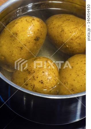 Close up view of several whole, unpeeled raw yellow potatoes soaking in cold water inside a metallic saucepan on a stovetop, preparing to be boiled. High quality photo Close up view of several whole, unpeeled raw yellow potatoes soaking in cold water inside a metallic saucepan on a stovetop, preparing to be boiled. High quality photo 140454430