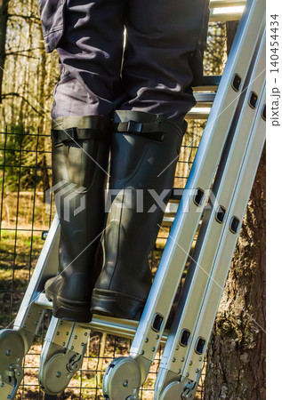 Person in rubber boots stands on an aluminum ladder for outdoor garden maintenance and home improvement projects in a sunny yard setting. High quality photo Person in rubber boots stands on an aluminum ladder for outdoor garden maintenance and home improvement projects in a sunny yard setting. High quality photo 140454434