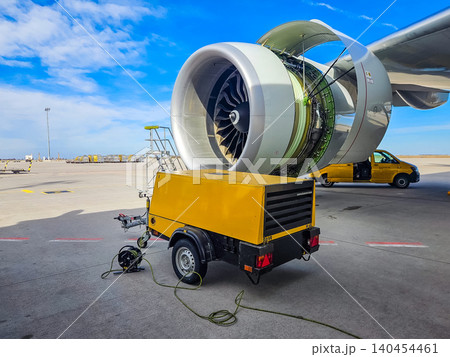 Close-up of an aircraft engine cowling open for inspection and repair with a mobile yellow power unit and service ladder at an airfield. High quality photo 140454461