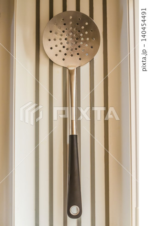 A well-used metal skimmer spoon, a common kitchen tool for straining or frying, hangs against a modern, clean, white grooved wall. High quality photo 140454491