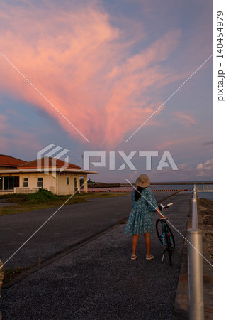 ピンク色の夕焼け雲を見上げる女性と自転車のある海辺の風景 140454979