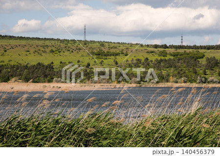 A wide summer landscape featuring a blue lake, lush green reeds, and rolling hills under a cloudy sky 140456379