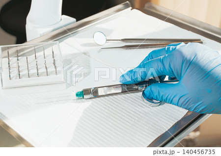 The dentist's hand, in a blue glove, holds a carpal syringe on a tray, ready for a medical procedure. The dentist's hand, in a blue glove, holds a carpal syringe on a tray, ready for a medical procedure. 140456735