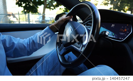 Male hand of businessman holds steering wheel while driving electrical vehicle at city. Businessperson operating electric car on urban road. Young man commuting to work at sunny day. Close up Male hand of businessman holds steering wheel while driving electrical vehicle at city. Businessperson operating electric car on urban road. Young man commuting to work at sunny day. Close up 140457247
