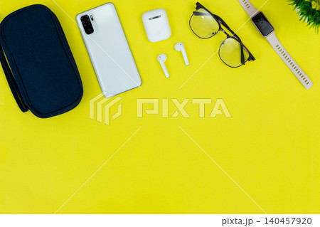 Bright yellow background holds tech and style. White phone rests near wireless earbuds and glasses. Laptop corner shows active workspace use. Bitcoin coin adds modern finance or digital theme 140457920