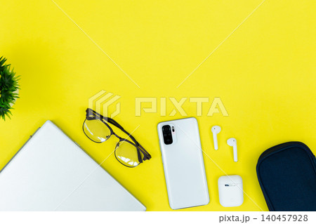 Bright yellow background holds tech and style. White phone rests near wireless earbuds and glasses. Laptop corner shows active workspace use. Bitcoin coin adds modern finance or digital theme Bright yellow background holds tech and style. White phone rests near wireless earbuds and glasses. Laptop corner shows active workspace use. Bitcoin coin adds modern finance or digital theme 140457928
